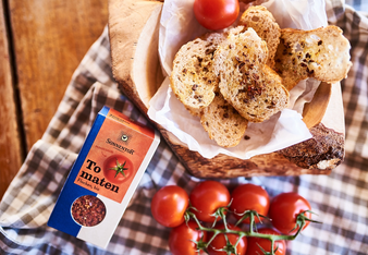 Das Päckchen Tomatenflocken liegt neben einem Korb knusprigen Baguette und einer Rispe frischer Cocktailparadeiser.  | © SONNENTOR