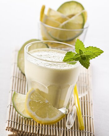 Auf dem Foto ist ein Glas mit Limoncello-Shake zu sehen. Davor sieht man Limetten und Zitronenscheiben. Das Glas ist mit Minze garniert. | © SONNENTOR