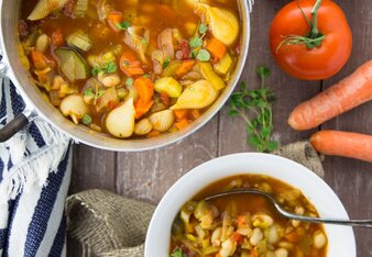 Au dem Foto sieht man einen Topf und einen Teller mit Minestrone. Daneben sieht man etwas Brot, Karotten und Tomaten. | © SONNENTOR