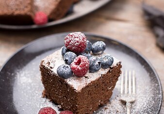 Auf dem Foto sieht man ein Stück Schoko Chai Kuchen. Er ist mit frischen Beeren und Staubzucker dekoriert. Im Hintergrund sieht man noch mehr Kuchenstücke. | © SONNENTOR