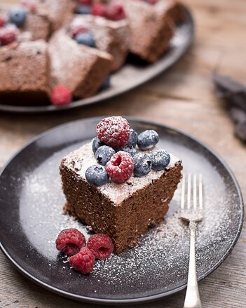 Auf dem Foto sieht man ein Stück Schoko Chai Kuchen. Er ist mit frischen Beeren und Staubzucker dekoriert. Im Hintergrund sieht man noch mehr Kuchenstücke. | © SONNENTOR