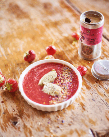 Auf dem Foto ist ein Holztisch zu sehen, auf dem ein weißes Schälchen mit Erdbeer-Melonen Suppe mit Flower Power als Topping. | © SONNENTOR