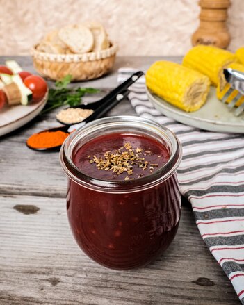 Die tiefrote BBQ-Sauce ist in einem bauchigen Glas, das zwischen Mais, einem Brotkörbchen mit Brot und Grillspießen auf dem Tisch steht. | © SONNENTOR