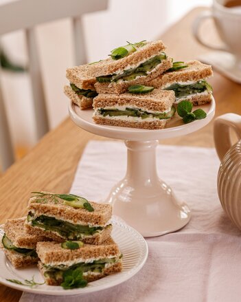 Einige Gurkensandwiches liegen auf einer weißen Etagere, einige auf einem Teller davor. Sie sind dekoriert mit einigen Gurkenscheiben und Minzblättern.  | © SONNENTOR