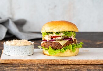 Der Cheeseburger ist gefüllt mit einem Rindfleischpattie, Salat, Paradeiser, Sauce, roten Zwiebeln und Käse und liegt auf einem Brett neben einem Schüsserl mit Sauce. | © SONNENTOR