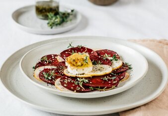 Auf dem Foto sieht man ein Teller mit rotem Beete Carpaccio. Darauf liegt Ziegenkäse. Im Hintergrund sieht man Gewürze und Kräuter. | © SONNENTOR