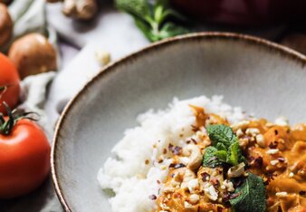 Auf dem Foto ist ein Teller mit Cashew-Curry zu sehen. Daneben ist auch Reis zu sehen. Im Hintergrund sieht man Ingwer und Tomaten. | © SONNENTOR