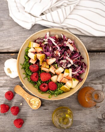 Ein Schüssel mit geschnittenem Rucola, Rotkraut und Birnen, mit Himbeeren am Rucola verteilt, steht zwischen einer Knoblauchknolle, einem Holzlöffel mit Knoblauchgranulat und 2 Dressingvasen auf einem Holztisch.  | © SONNENTOR