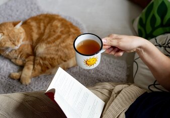 Auf dem Foto trinkt eine Frau auf dem Sofa eine Tasse Tee und liest ein Buch. Daneben sieht man eine Katze liegen. | © SONNENTOR