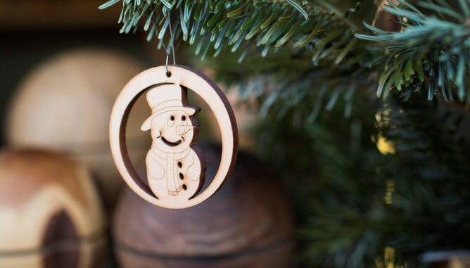 Auf dem Foto sieht man einen Christbaumaufhänger aus Holz mit einem Schneemannmotiv. Das Dekostück hängt auf einer Tanne. | © SONNENTOR